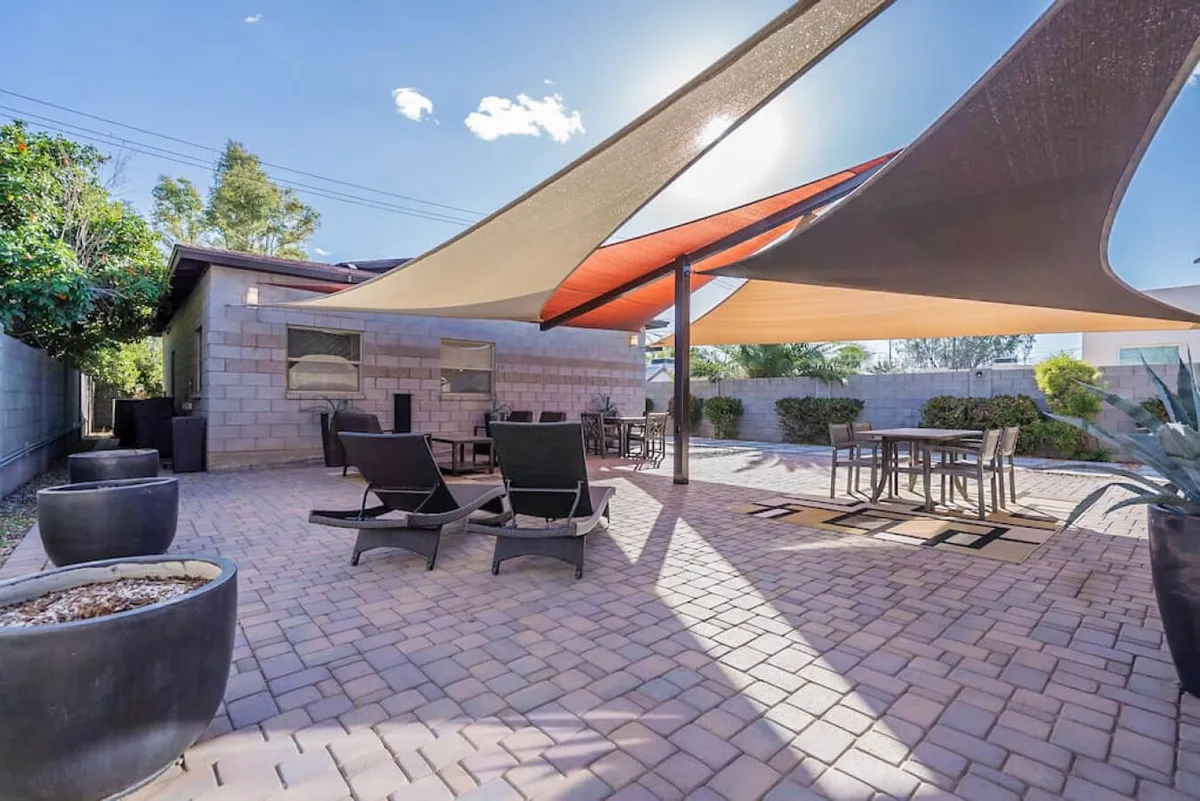 This exterior image captures a shaded patio area featuring modern shade sails, inviting patio furniture, and lush landscaping. The scene suggests a tranquil, welcoming space ideal for outdoor relaxation and casual gatherings, nestled within a leafy, residential environment.