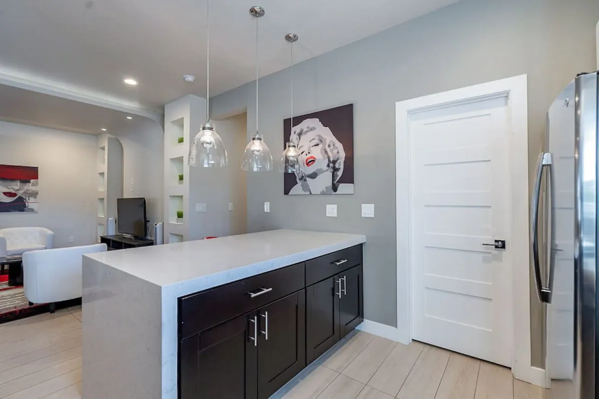 A modern kitchen with a spacious island countertop, stylish pendant lights, and iconic Marilyn Monroe artwork, inviting interaction and group activity. The kitchen's open layout and contemporary decor suggest a welcoming space for shared meals and lively gatherings after a day of exploring Phoenix.