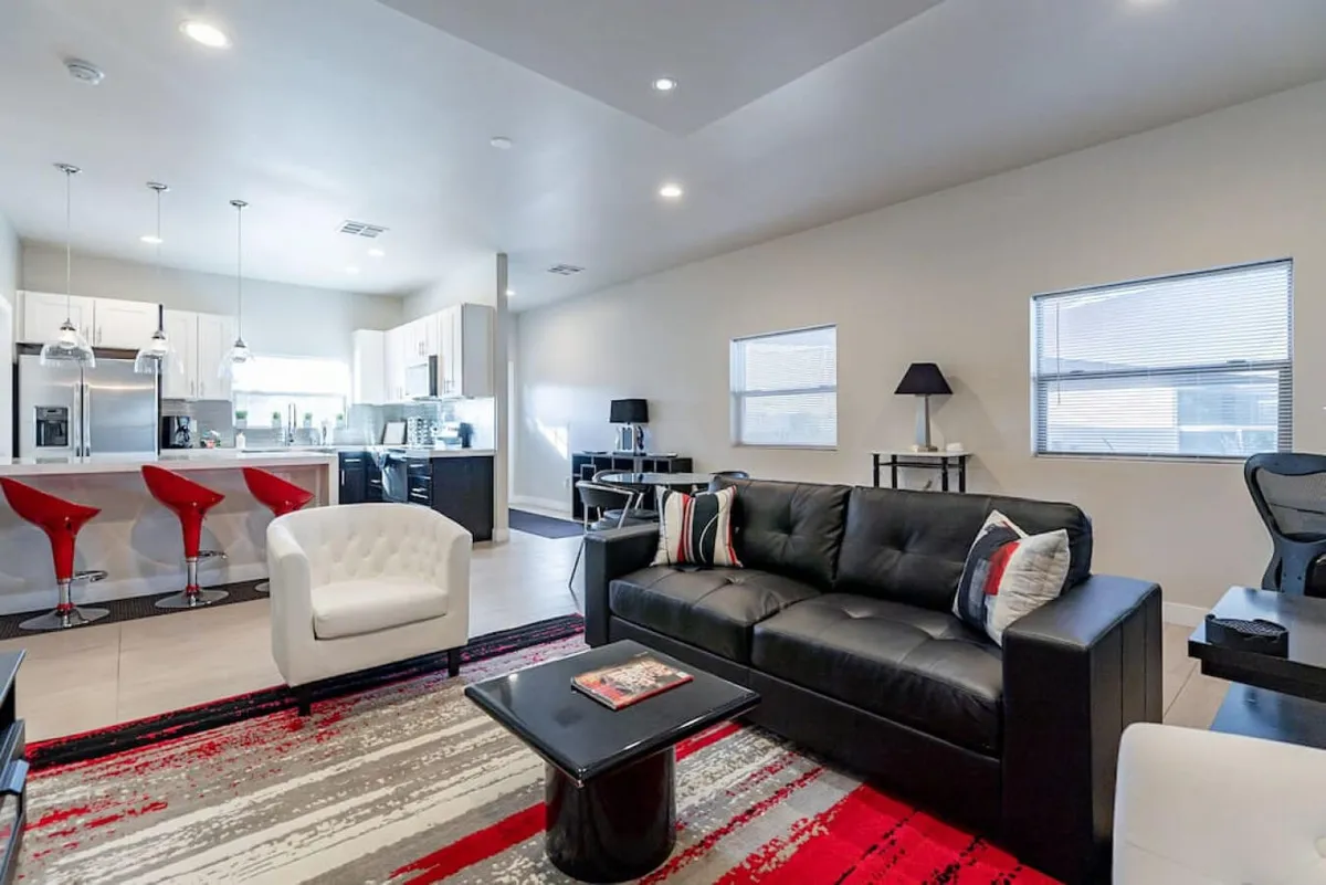 A vibrant living room anchored by a black leather sofa, red bar stools, white armchair, and a colorful area rug. The room offers a lively, welcoming atmosphere ideal for socializing, relaxing, or enjoying various activities in comfort and style.