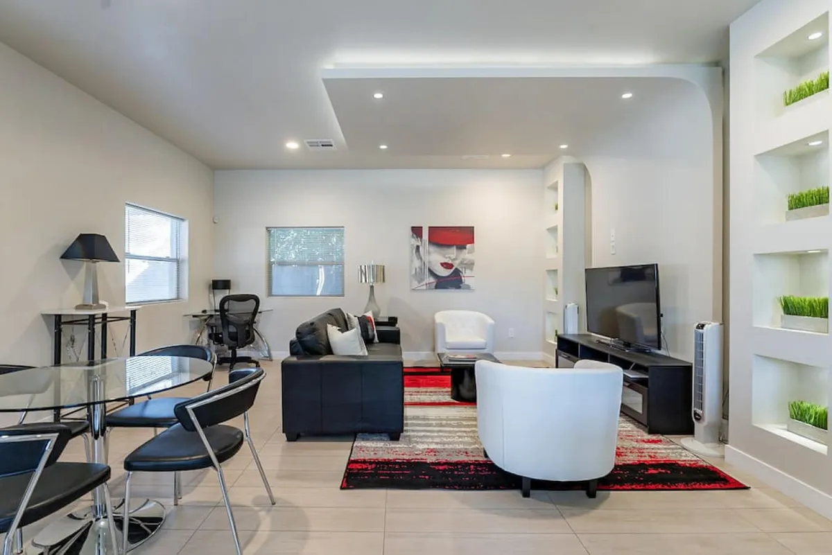 A bright and cozy living/dining area featuring a black leather sofa, white accent chair, glass dining table, TV unit, and vibrant artwork. The inviting arrangement is ideal for group relaxation, watching TV, or recounting the day's adventures.