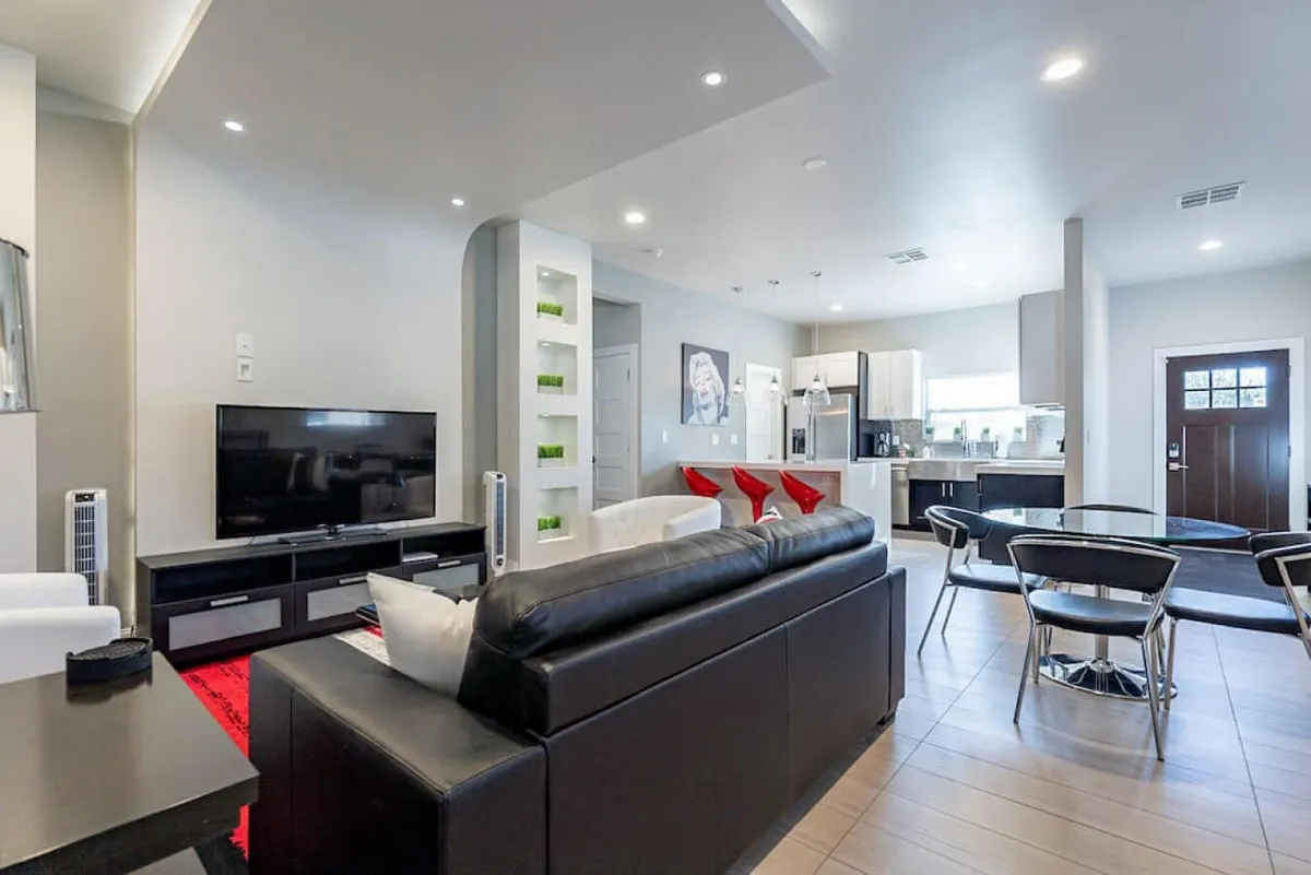 A welcoming open-plan living and dining area, featuring a glass dining table with chrome chairs adjacent to kitchen appliances, and a cozy black leather sofa with red accent pillows. The space is arranged for easy entertaining and comfortable dining, with natural flow between cooking, eating, and socializing.