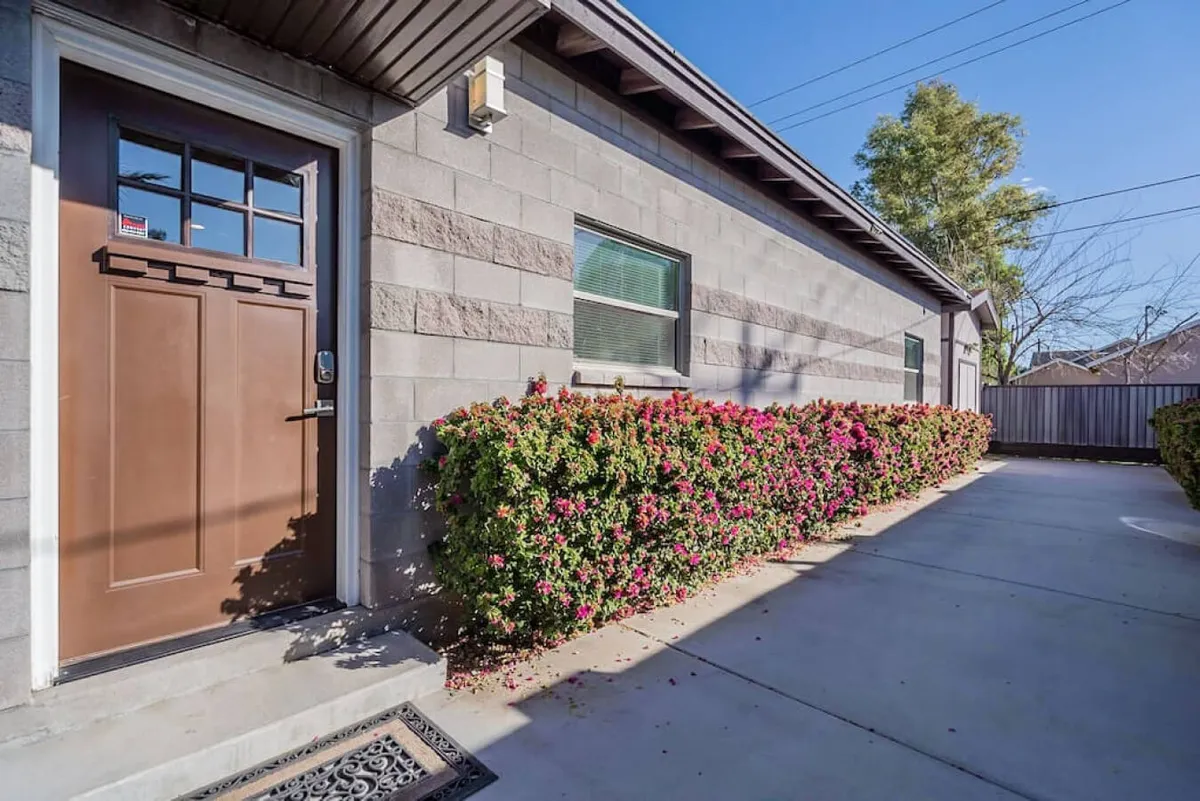 A welcoming view of the property’s front door framed by flowering bushes, a clean sidewalk, and a tidy exterior wall. The mood is inviting and gives a strong sense of arrival at a quiet residential base.