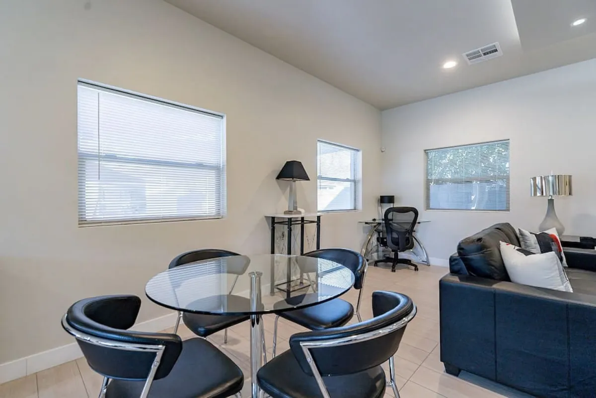 The image presents a contemporary, inviting interior living space with a glass dining table, a sleek black leather sofa, a work desk with a chair, and warm lighting from a lampshade. The arrangement suggests flexible usage for dining, working, or relaxing.