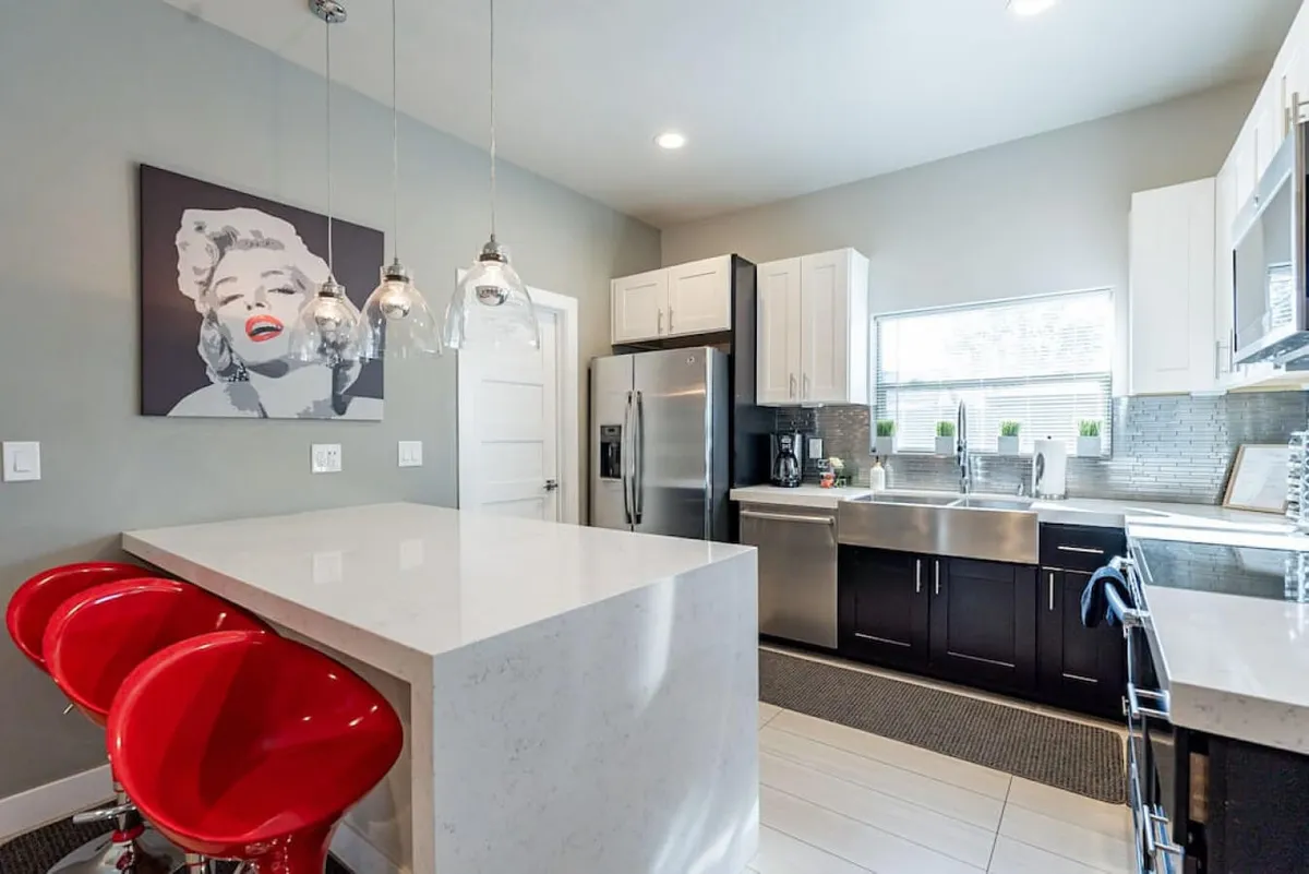 A vibrant kitchen scene with striking red bar stools at a white marble countertop, stainless steel appliances, and a bold Marilyn Monroe artwork. The setup highlights both functionality and style, providing a lively space for casual dining or cooking with friends.