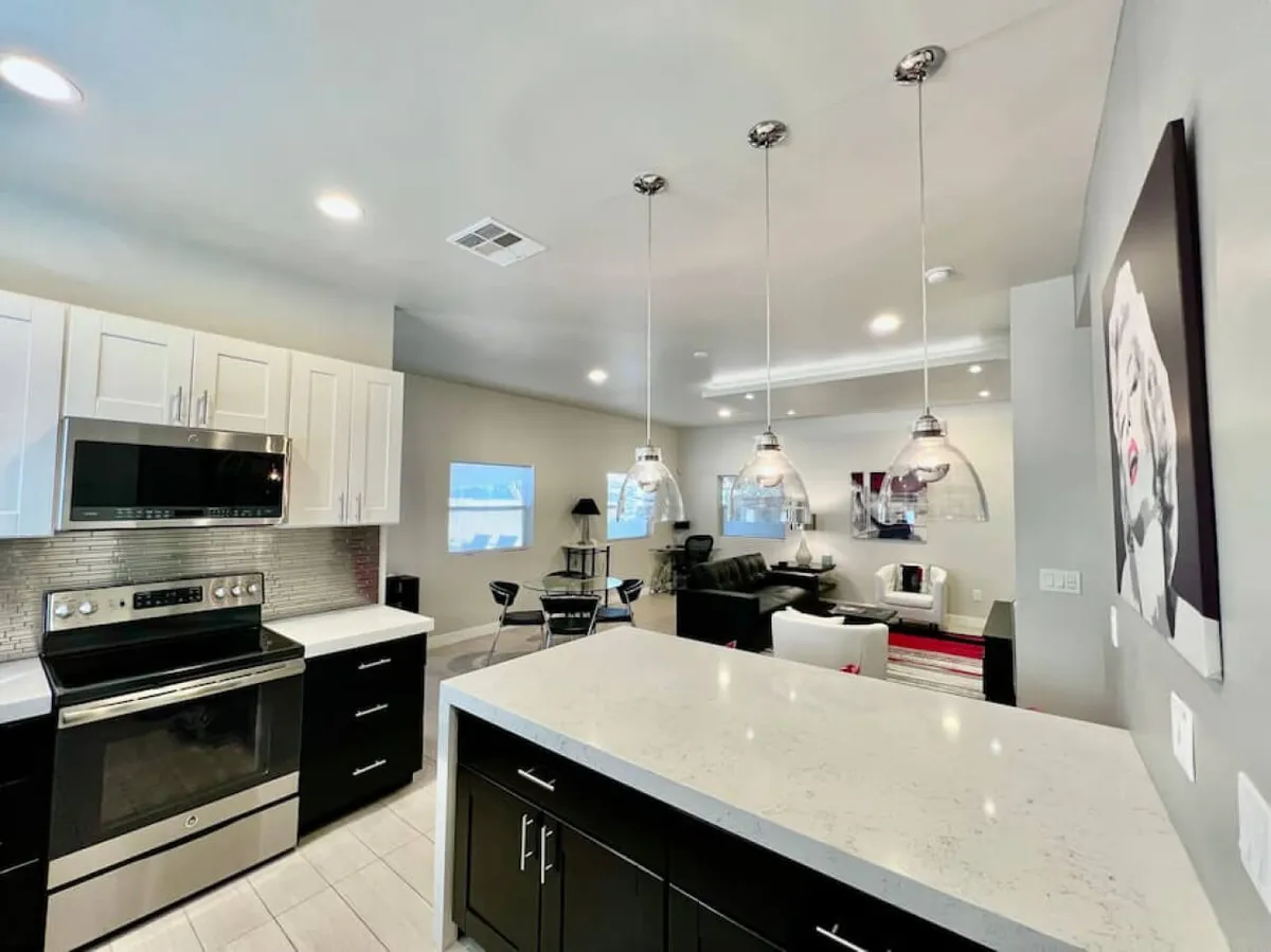 The image showcases a bright, modern open-concept kitchen and living area. A spacious kitchen island with stylish pendant lighting takes center stage, surrounded by comfortable seating and accented by vibrant artwork. The scene feels inviting, perfect for hosting, relaxing, and gathering with loved ones.