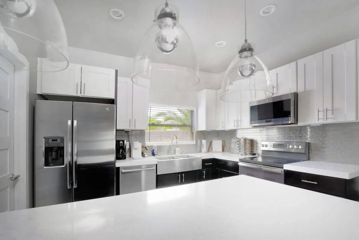 A bright, contemporary kitchen featuring a central island with white cabinetry, stainless steel appliances, and hanging pendant lights. The layout offers an open space ideal for both meal prep and social gatherings, with ample room around the island for group conversations or family activities.