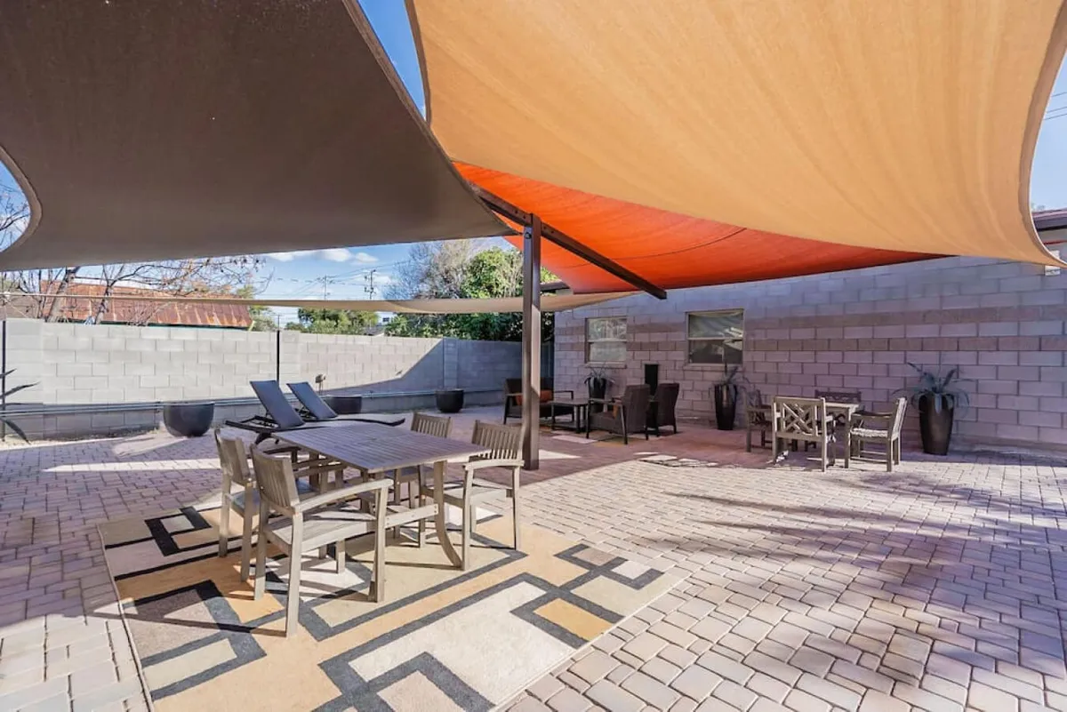 An outdoor social space with a dining table, lounge chairs, shade sails, and decorative planters. The setup suggests gatherings, outdoor meals, and relaxed afternoons just outside the home.