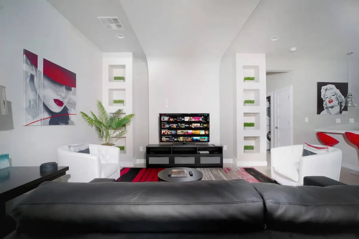 This interior image features a contemporary living space with a black couch, stylish artwork, a large television, and a green plant adding a touch of freshness. The arrangement emphasizes a relaxing and welcoming atmosphere, with natural light filtering in and modern décor.